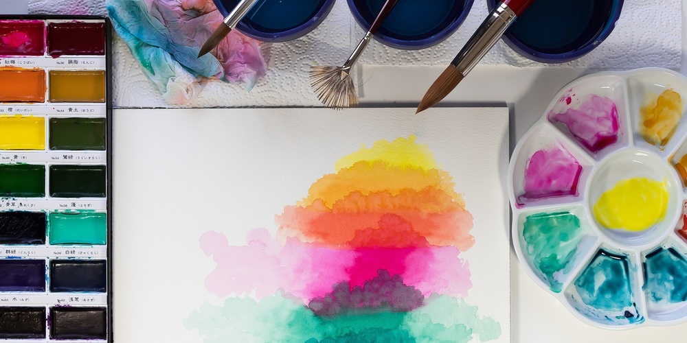 Ett akvarellmålningsset med färger, penslar och en målning i regnbågens färger på ett vitt papper.