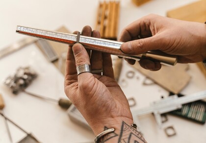 Två händer håller ett ringmåttverktyg ovanför ett bord med smyckestillverkningsmaterial.