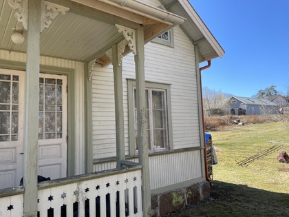 Gammalt trähus med veranda och snickarglädje, solig dag med gräsmatta och andra hus i bakgrunden.