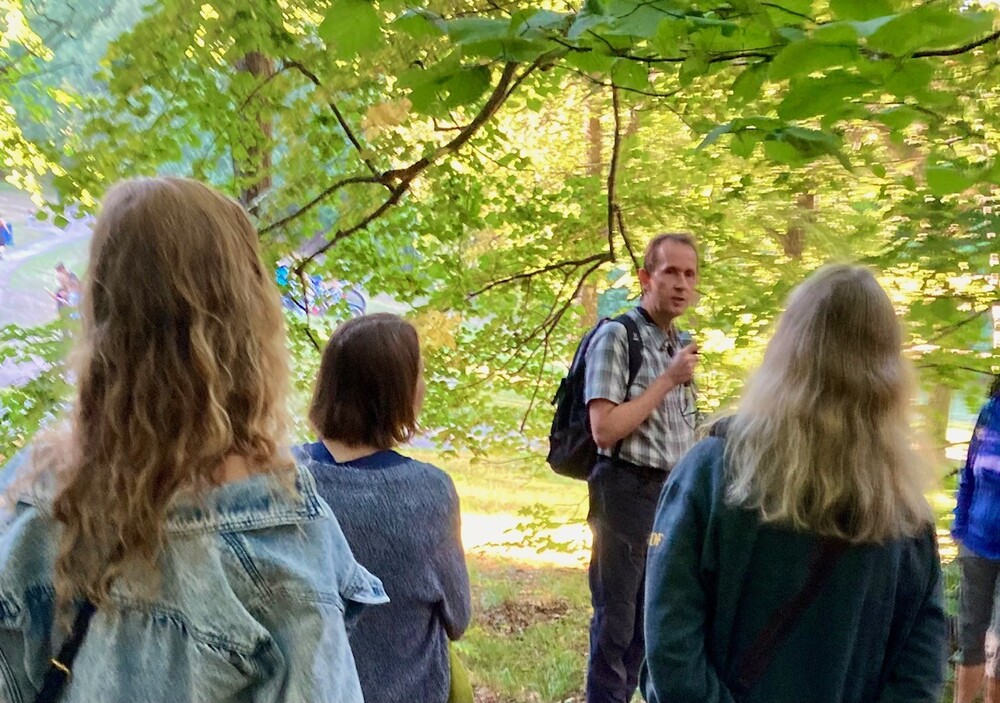 En grupp människor står utomhus i en grönskande park, en man pratar till gruppen med mikrofon.