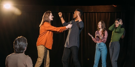 Fyra personer står på en scen och spelar teater, medan en person sitter och tittar på dem.