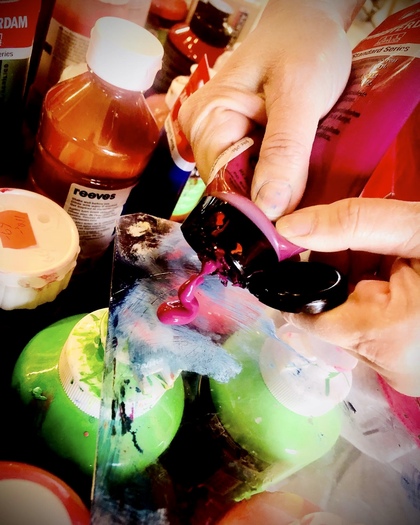 Närbild av händer som klämmer ut rosa akrylfärg på en palett bland färgtuber och burkar.