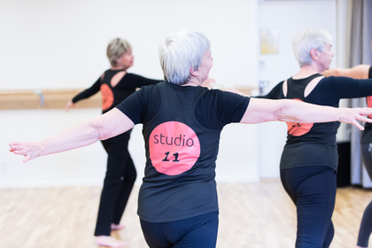Äldre kvinnor deltar i en dans- eller träningsklass, iförda svarta kläder med texten 'studio 11' på ryggen.