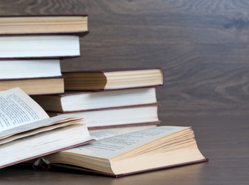 Flera böcker ligger och staplas på ett träbord, några är öppna och andra stängda.