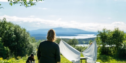 En person står på en gräsmatta och tittar mot ett tält av vita lakan, med fler personer i bakgrunden och sjöutsikt.