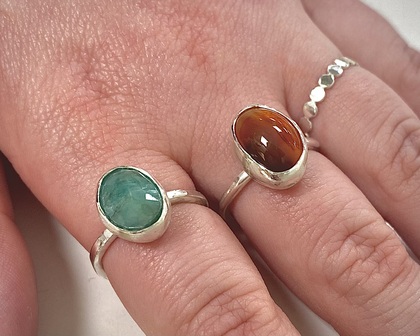 Två händer med silverringar, en med grön sten och en med brun sten, samt en tunn ring med små cirklar.