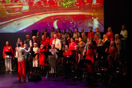 En kör med vuxna sjunger på en scen med färgglad bakgrundsbild och dirigent framför dem.