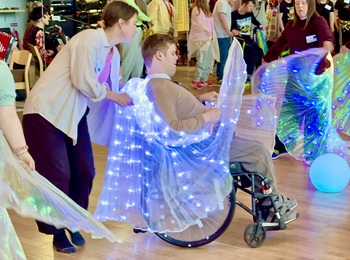 # personer i ett rum varav en sitter i en rullstol. De dansar med färggladda isisvingar. En fjärde person hjälper personen i rullstolen