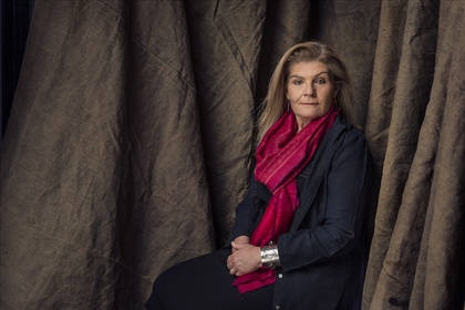 Kvinna med långt hår och röd sjal sitter framför bruna tygdraperier och tittar mot kameran.