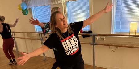 Kvinna dansar glatt i en dansstudio med trägolv och speglar, iförd svart t-shirt med texten 'DANS STUDIO'.