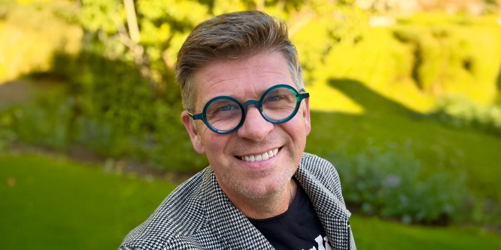 Person i rutigt kavaj och svart t-shirt står utomhus i en grönskande park.