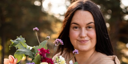 Person håller en stor bukett färgglada blommor utomhus i naturligt ljus.
