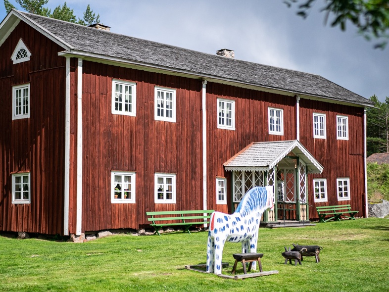 Röd trähus med vita fönster, gröna bänkar och en vitblå prickig häststaty på gräsmattan framför huset.