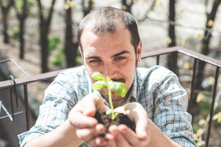 En man håller en liten planta med jord i händerna och ler mot kameran utomhus.