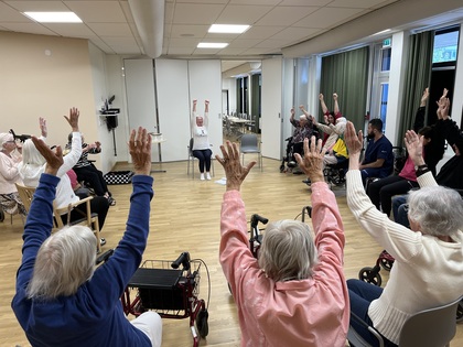 En grupp äldre personer sitter i en cirkel och gör sittande gymnastik med armarna uppsträckta.