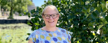 Kvinna med glasögon och mönstrad klänning står utomhus vid ett träd en solig dag.