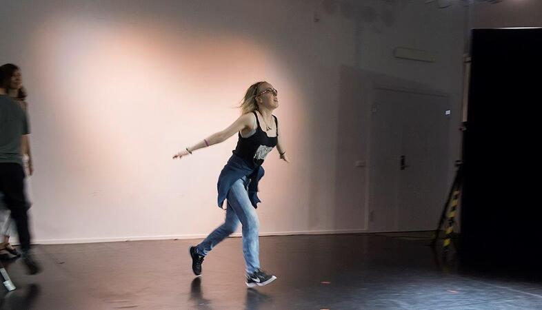 En person springer eller dansar på en scen i ett rum med ljusa väggar och mörkt golv.