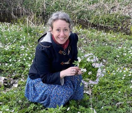 Person i blå kjol plockar blommor på en äng med vita och lila blommor nära en bäck.