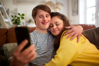 Två personer sitter i en soffa och tar en selfie tillsammans, de kramar om varandra och ser glada ut.