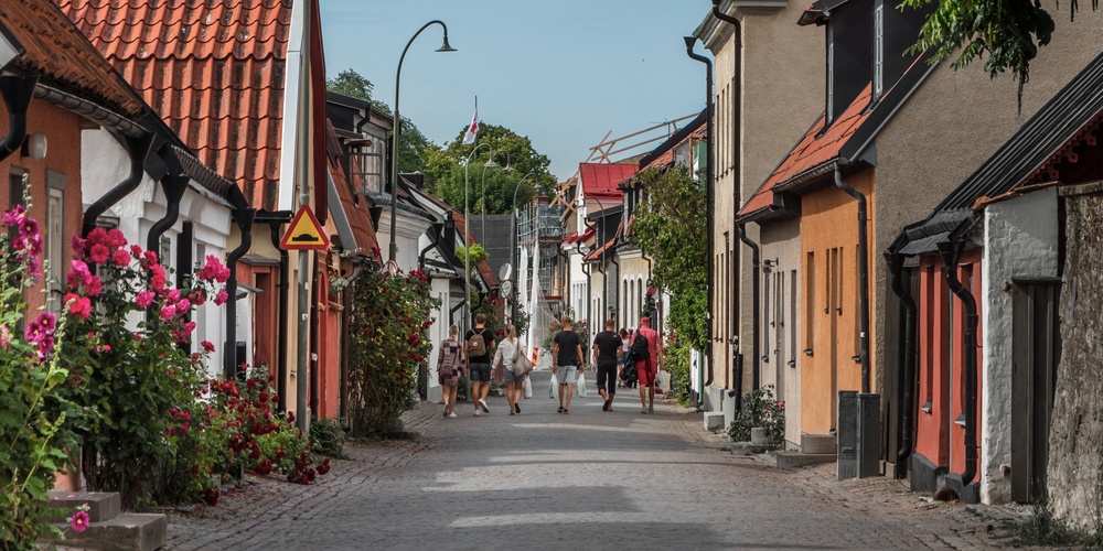 En pittoresk kullerstensgata med färgglada hus, blommor och människor som promenerar en solig dag.