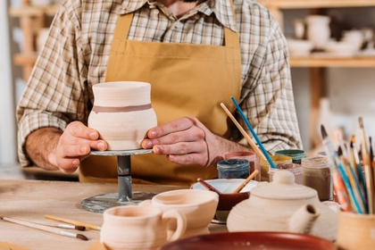 En person i förkläde arbetar med keramik på en drejskiva i en verkstad fylld med lergods och penslar.