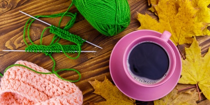 Stickning med grönt garn, rosa stickat plagg, rosa kaffekopp och gula höstlöv på ett träbord.