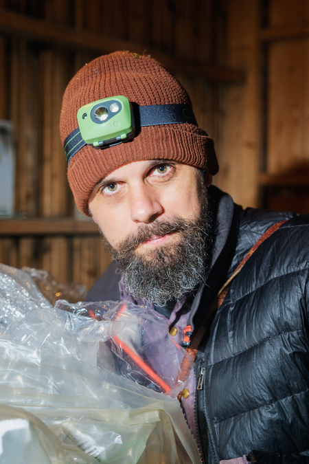 Person med orange stickad mössa och svart jacka håller i plastpåsar i en träbyggnad.