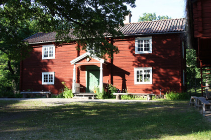 Röd trähus med vita fönster och grön dörr, omgiven av träd och gräsmatta i solsken.