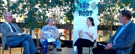 Fyra personer sitter i en paneldiskussion inomhus med gröna växter och en banner i bakgrunden.