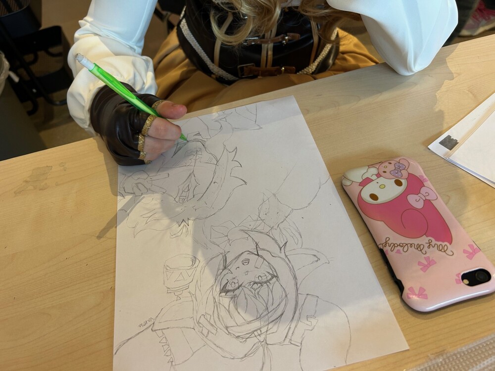 Person tecknar animefigurer på ett papper vid ett bord, med en mobiltelefon med rosa fodral bredvid.