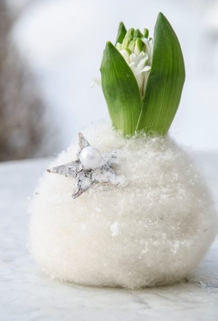 En grön växt med vita blommor växer ur en boll täckt av konstsnö, dekorerad med en silverstjärna och pärla.