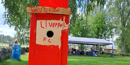 En röd fågelholk med texten 'Liverpool FC' står på ett träbord utomhus med grönskande träd i bakgrunden.