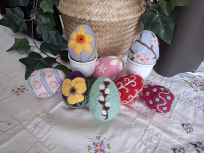 Färgglada tovade påskägg med blommönster ligger på en duk med broderade blommor, omgivna av gröna växter.