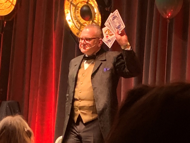 En man i kostym står på en scen och håller upp stora spelkort inför en publik.