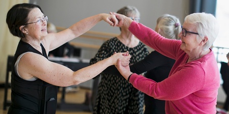 Två äldre kvinnor dansar tillsammans i en ljus dansstudio med andra personer i bakgrunden.