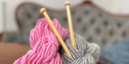 Två garnnystan, ett rosa och ett grått, med stickor på en färgglad virkad filt.