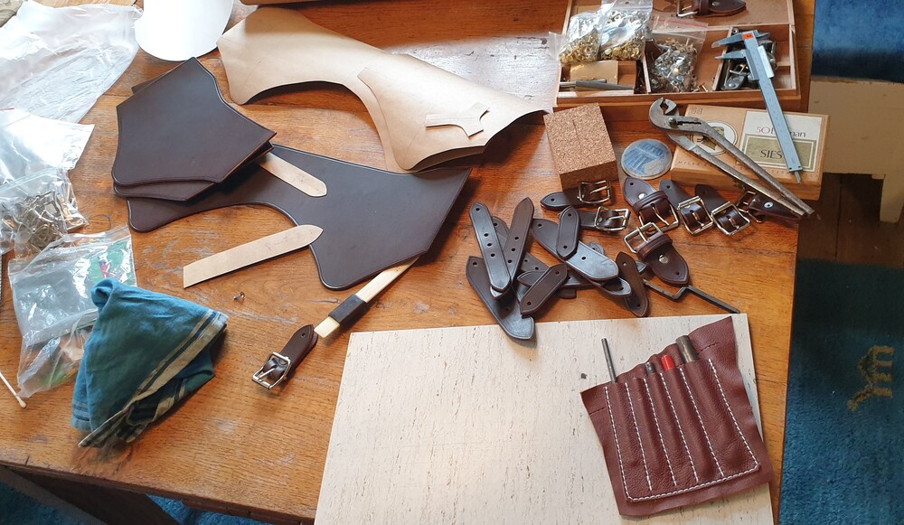 Ett arbetsbord med verktyg och material för läderhantverk, inklusive spännen, läderbitar och verktyg.