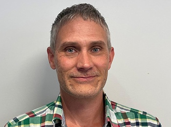 Person med kort hår och rutig skjorta står mot en ljus vägg.