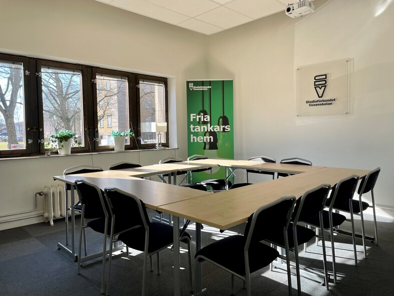 Ljust konferensrum med U-format bord, svarta stolar och stora fönster. Grön skylt med texten 'Fria tankars hem'.