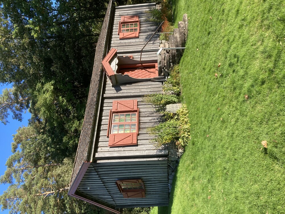 En liten grå trästuga med röda fönsterluckor och dörr, omgiven av grön gräsmatta och träd.