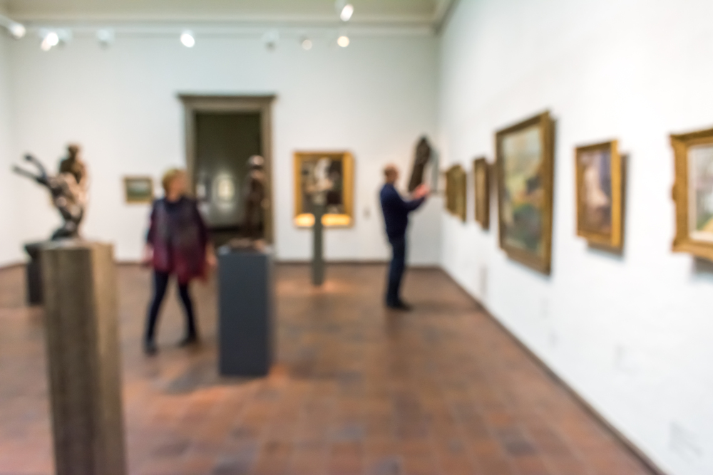 Suddig bild av personer som tittar på konstverk i ett galleri med tavlor och skulpturer.