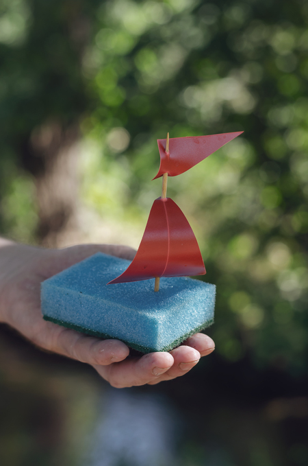 En hand håller en liten båt gjord av en blå tvättsvamp, en träpinne och röda segel utomhus.