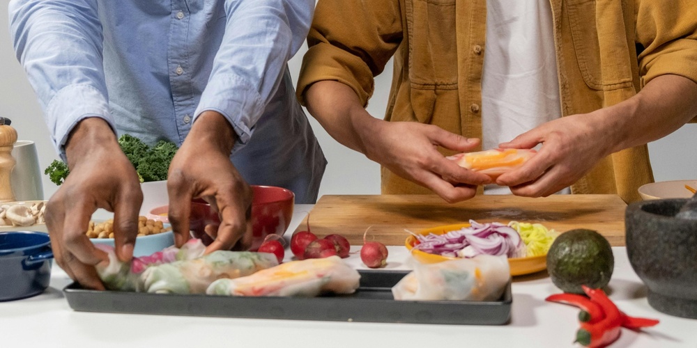 Två personer förbereder färska vårrullar med grönsaker på ett köksbord.