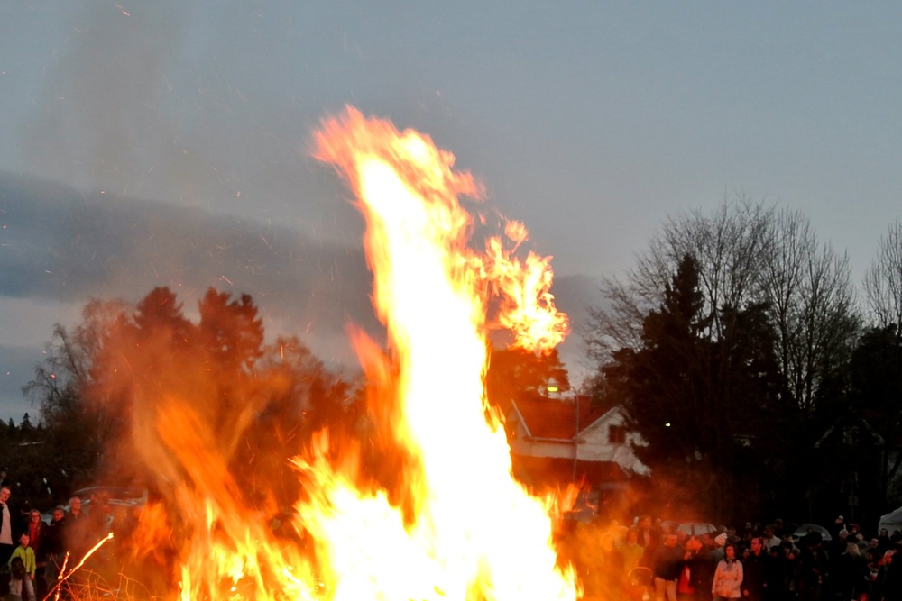 Stor brasa brinner på en gräsmatta omgiven av människor under kvällstid.