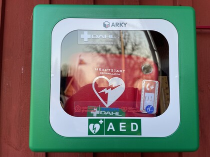 Grön förvaringsbox för hjärtstartare (AED) monterad på en röd vägg.
