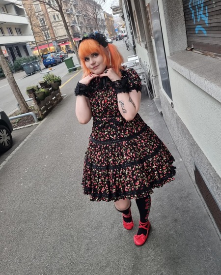Person med orange hår och svart blommig klänning poserar på trottoar i stadsmiljö.