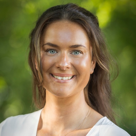 Kvinna med långt brunt hår och vit blus ler mot kameran utomhus med grönskande bakgrund.