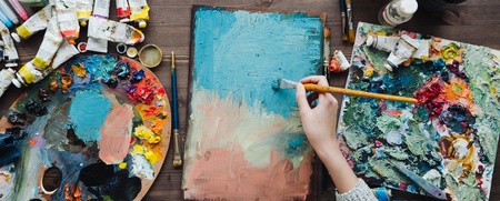 En person målar med pensel på en duk omgiven av färgtuber, penslar och paletter på ett träbord.