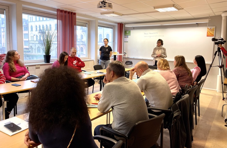 En grupp vuxna sitter runt ett bord i ett klassrum och lyssnar på en presentation vid whiteboard.