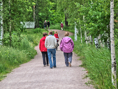 Flera personer promenerar på en grusväg omgiven av gröna träd och gräs en sommardag.